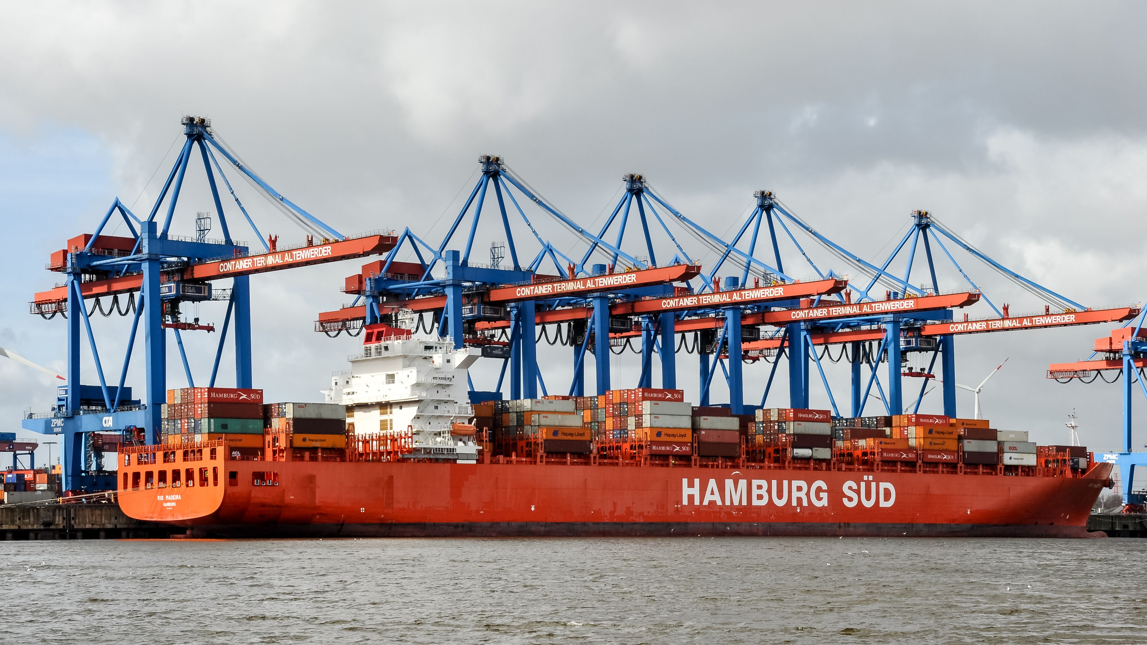 Red container ship docked at Hamburg Terminal under cranes on a cloudy day.